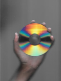 A hand holding a compact disc, which displays a spectrum of colors ranging from bright red, through orange, yellow, green, and blue, against a gray background.