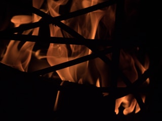 Dancing flames spread across a dark background, with silhouetted intersecting lines in the foreground. The light and shadow create a striking contrast, highlighting the fluidity and intensity of the fire.