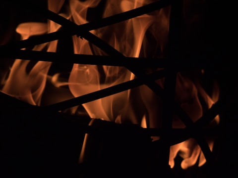 Dancing flames spread across a dark background, with silhouetted intersecting lines in the foreground. The light and shadow create a striking contrast, highlighting the fluidity and intensity of the fire.