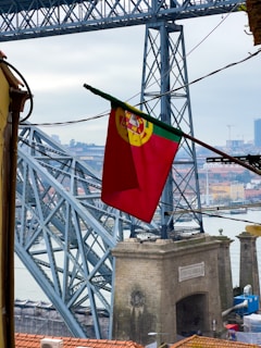 A large iron bridge dominates the scene with intricately interwoven beams and supports. In the foreground, a vibrant red flag with a yellow and green emblem flutters gently. Below the bridge, there's a stone archway labeled with the words 'Ponte Luiz I' while the cityscape stretches in the background, revealing various buildings and the river.