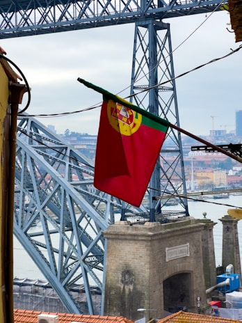 A large iron bridge dominates the scene with intricately interwoven beams and supports. In the foreground, a vibrant red flag with a yellow and green emblem flutters gently. Below the bridge, there's a stone archway labeled with the words 'Ponte Luiz I' while the cityscape stretches in the background, revealing various buildings and the river.