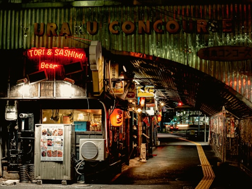 A dimly lit urban street scene features small eateries and bars under a metal-covered concourse. Neon signs, including one for 'Tori & Sashimi' and 'Beer,' illuminate the otherwise dark area. The alleyway is narrow, with various posters and signs on the walls and a visible air conditioning unit.