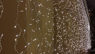 A dense array of warm white LED string lights is draped vertically, creating a shimmering curtain effect against a tan background. The lights form intricate patterns due to their intertwining cords, adding depth and texture to the scene.