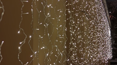 A dense array of warm white LED string lights is draped vertically, creating a shimmering curtain effect against a tan background. The lights form intricate patterns due to their intertwining cords, adding depth and texture to the scene.