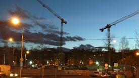 Cranes tower over a construction site at dusk, with streetlights illuminating the area and casting a warm glow on the scene. The sky is a mix of blue and dark clouds, suggesting an impending nightfall. Bare trees and a few buildings are visible in the background, and traffic signs are present in the foreground.