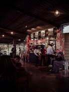 Interior of a coffee shop or restaurant with a counter in the middle background, adorned with red patterned decorations. Several people are interacting around the counter, some are staff members wearing uniforms and headscarves. There are hanging light fixtures and a series of photographs or artwork displayed above the counter. In the left foreground, a person is sitting at a table, looking down, possibly using a phone. On the right, people are sitting on stools or chairs, one person looking at a device.