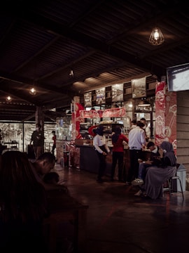 Interior of a coffee shop or restaurant with a counter in the middle background, adorned with red patterned decorations. Several people are interacting around the counter, some are staff members wearing uniforms and headscarves. There are hanging light fixtures and a series of photographs or artwork displayed above the counter. In the left foreground, a person is sitting at a table, looking down, possibly using a phone. On the right, people are sitting on stools or chairs, one person looking at a device.