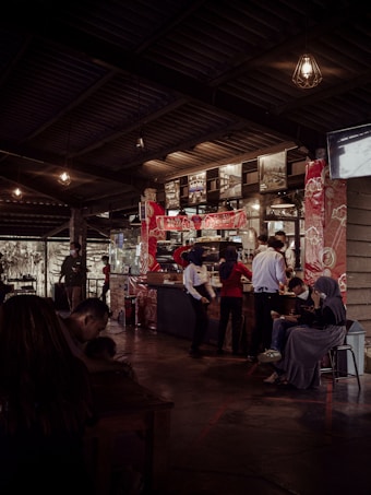Interior of a coffee shop or restaurant with a counter in the middle background, adorned with red patterned decorations. Several people are interacting around the counter, some are staff members wearing uniforms and headscarves. There are hanging light fixtures and a series of photographs or artwork displayed above the counter. In the left foreground, a person is sitting at a table, looking down, possibly using a phone. On the right, people are sitting on stools or chairs, one person looking at a device.