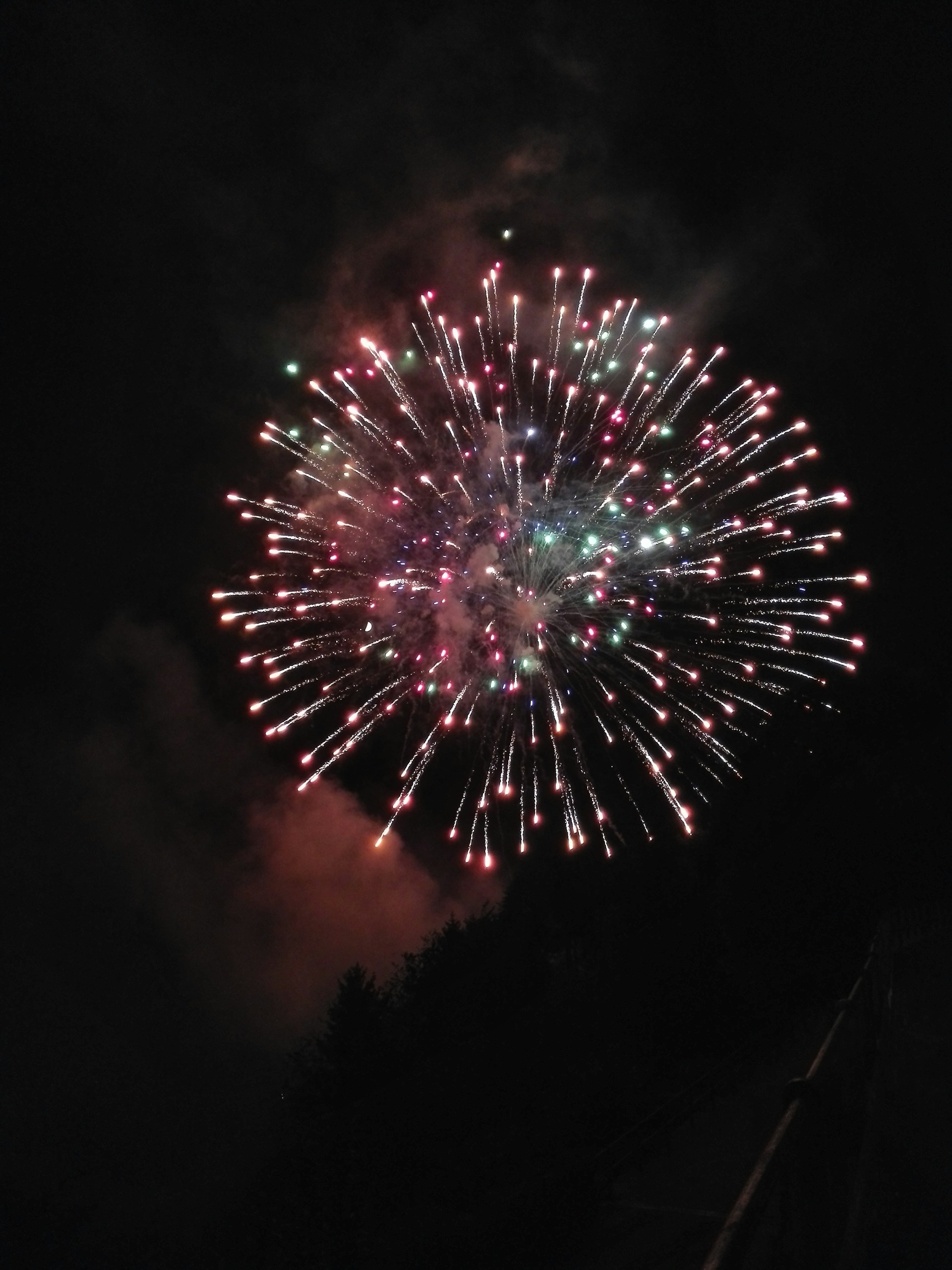 A vibrant burst of fireworks lights up the night sky with colorful streaks and sparks radiating outward. The explosion takes on a spherical shape, with a mix of bright colors including red, green, and pink. The contrast against the dark sky enhances the luminous effect of the fireworks.