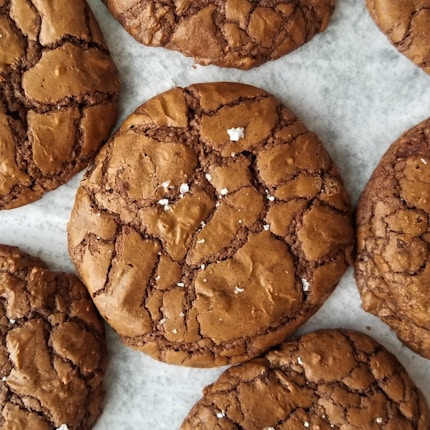 Several large, brown, crackly cookies arranged on a marble surface. The cookies have a glossy appearance with a few white specks on top.