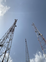 Several tall communication towers stretch upwards towards a clear blue sky. The metal structures are composed of interlocking beams and feature various equipment attached to them. Fluffy white clouds are scattered across the sky, adding contrast to the scene.