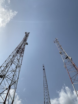 Several tall communication towers stretch upwards towards a clear blue sky. The metal structures are composed of interlocking beams and feature various equipment attached to them. Fluffy white clouds are scattered across the sky, adding contrast to the scene.