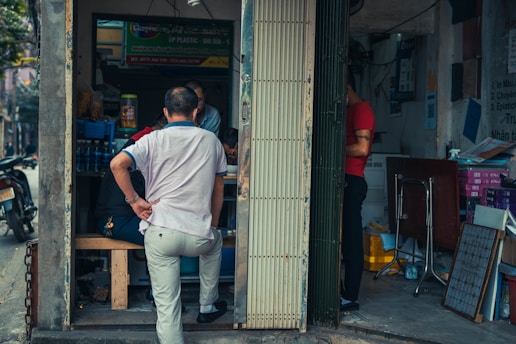 A small, dimly lit shop with several people inside and one person standing at the entrance. The shop is cluttered with various items including bottles, boxes, and a small table with chairs. A motorbike is partially visible outside the entrance.