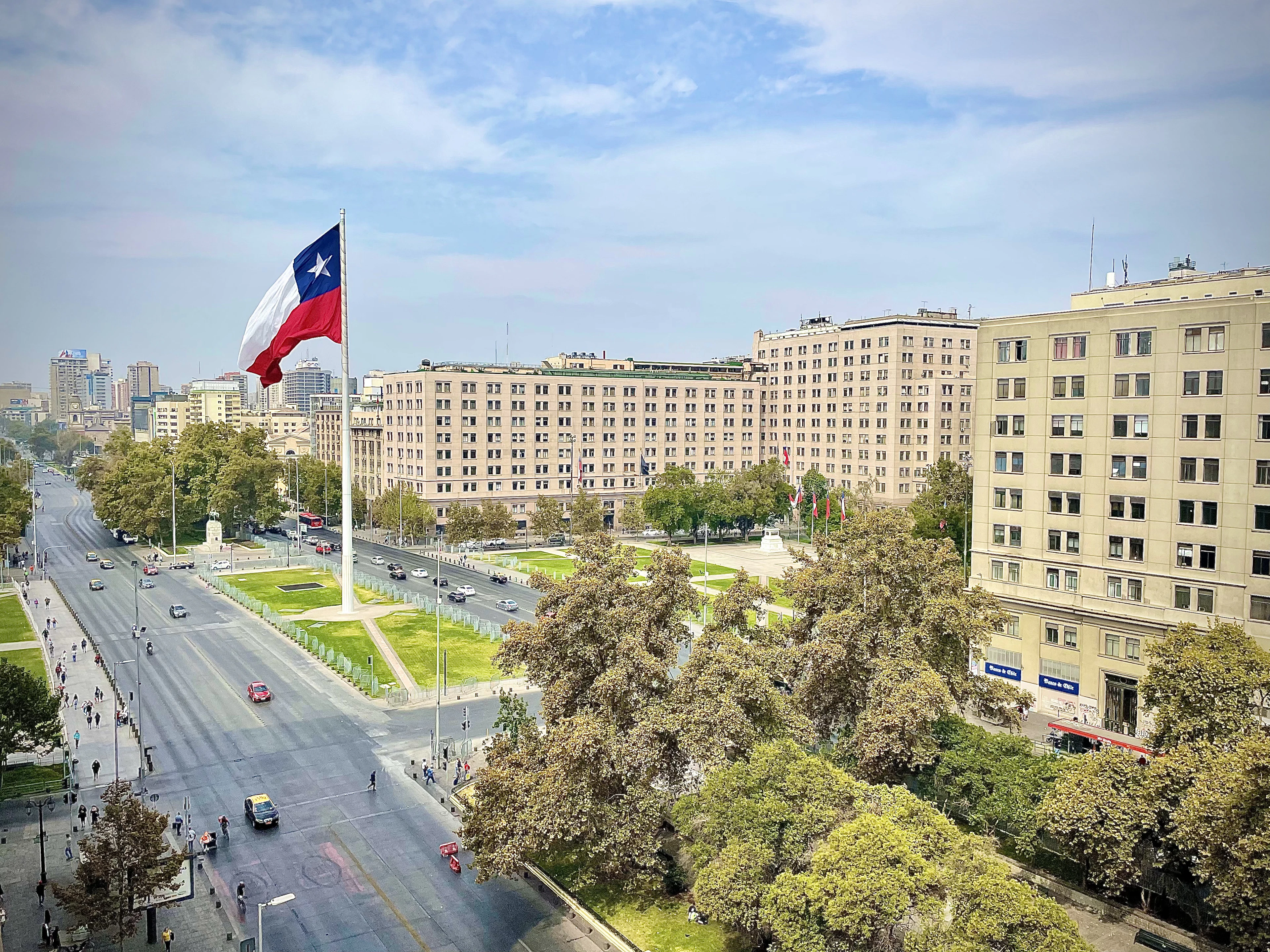 A giant Chilean flag flies over a broad Santiago boulevard lined with historic buildings and trees.