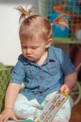 A young child with blonde hair in pigtails wearing a blue denim shirt and light-colored pants is intently looking at a book with colorful illustrations. The child is seated on the floor next to a green woven basket. In the background, there are some colorful toys on a wire shelf.