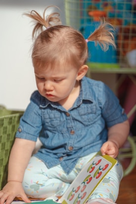A young child with blonde hair in pigtails wearing a blue denim shirt and light-colored pants is intently looking at a book with colorful illustrations. The child is seated on the floor next to a green woven basket. In the background, there are some colorful toys on a wire shelf.