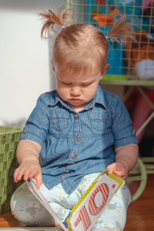 A young child with light hair styled in pigtails is sitting on the floor, concentrating on an open book in her hands. She is wearing a denim shirt and patterned leggings. The child appears to be deeply engaged in the activity of reading. In the background, there is a wire shelf with various colorful objects.