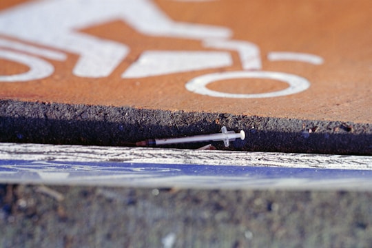 A discarded syringe is placed on the ground against the edge of a paved surface. The area features a painted bicycle symbol, indicating it may be near a bike path. The syringe appears partially hidden beneath the edge.