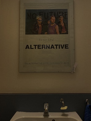 A poster is displayed above a sink in a bathroom. The poster features three individuals with punk style clothing and hairstyles. The text on the poster includes 'No Future' at the top and 'Es gibt EINE ALTERNATIVE' below, along with 'Die ALTERNATIVE LISTE ÖSTERREICH (ALÖ)'. A handwritten note is taped to the side of the poster.