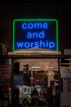 A brightly illuminated neon sign displays the words 'come and worship' in blue lettering, bordered by a green outline. The sign is part of a storefront with reflective glass showing a mix of posters and signs, including a notice about wearing face coverings. The interior appears to have warm lighting with reflections capturing street traffic and passersby.