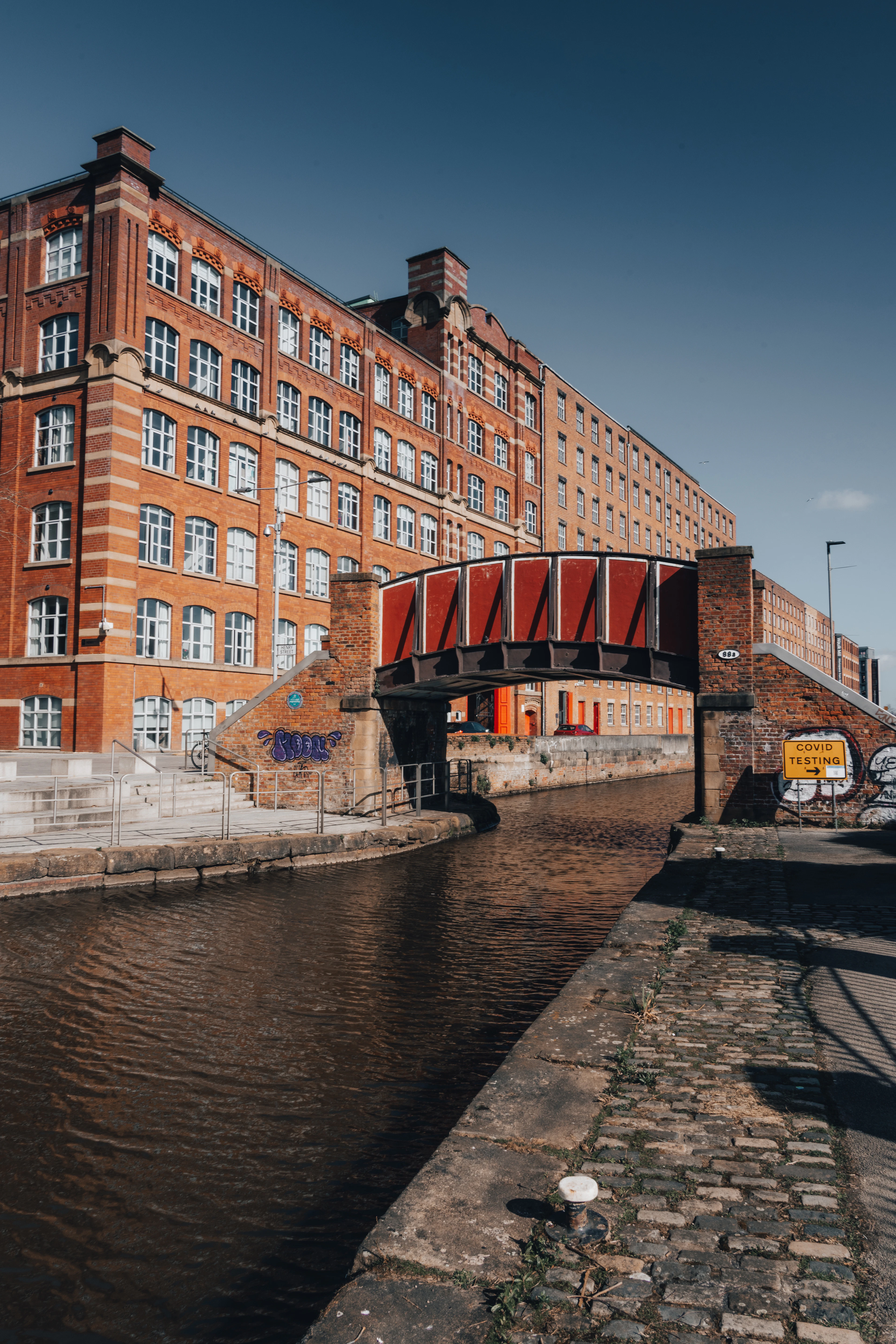 A tall red-brick cotton mill standing over the canal in Manchester's Northern Quarter, a red iron footbridge crossing the water in low sun.