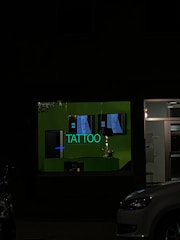 A tattoo shop window is dimly illuminated by a green neon sign displaying the word 'TATTOO'. There are posters inside showing tattoo designs, and some of the shop's interior can be seen through the glass. A car is partially visible parked outside.
