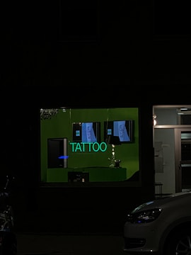 A tattoo shop window is dimly illuminated by a green neon sign displaying the word 'TATTOO'. There are posters inside showing tattoo designs, and some of the shop's interior can be seen through the glass. A car is partially visible parked outside.