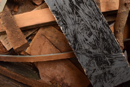 A collection of wooden planks and plywood pieces, arranged in a haphazard manner. The textures of the wood vary, with one prominently featuring dark, intricate patterns typical of oriented strand board. The colors range from light brown to dark brown, with some areas appearing worn and rough.