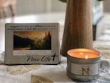A decorative plaque with a motivational Christian message is propped up next to a lit nutmeg-scented candle on a patterned tablecloth. The plaque features a photograph of a serene landscape with mountains and trees during sunset. The overall setting is cozy and calm.
