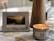 A decorative plaque with a motivational Christian message is propped up next to a lit nutmeg-scented candle on a patterned tablecloth. The plaque features a photograph of a serene landscape with mountains and trees during sunset. The overall setting is cozy and calm.
