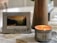 A decorative plaque with a motivational Christian message is propped up next to a lit nutmeg-scented candle on a patterned tablecloth. The plaque features a photograph of a serene landscape with mountains and trees during sunset. The overall setting is cozy and calm.
