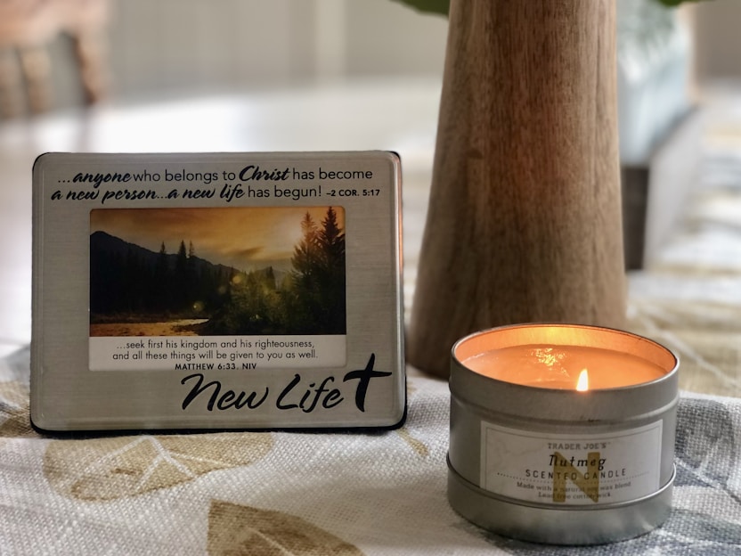 A decorative plaque with a motivational Christian message is propped up next to a lit nutmeg-scented candle on a patterned tablecloth. The plaque features a photograph of a serene landscape with mountains and trees during sunset. The overall setting is cozy and calm.