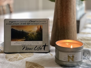 A decorative plaque with a motivational Christian message is propped up next to a lit nutmeg-scented candle on a patterned tablecloth. The plaque features a photograph of a serene landscape with mountains and trees during sunset. The overall setting is cozy and calm.