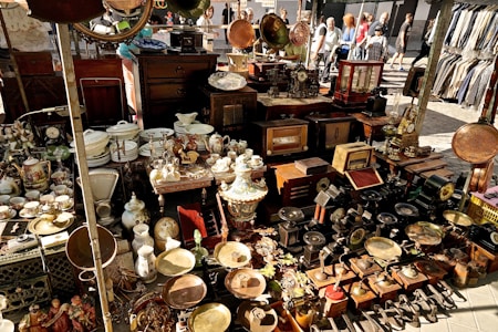 A bustling marketplace stall displays a variety of vintage and antique items, including ornate clocks, porcelain dishes, decorative vases, old radios, and brass objects. The items are densely packed on tables and shelves. In the background, people are walking past the stall, creating a lively outdoor market atmosphere.