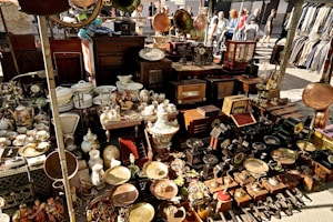 A bustling marketplace stall displays a variety of vintage and antique items, including ornate clocks, porcelain dishes, decorative vases, old radios, and brass objects. The items are densely packed on tables and shelves. In the background, people are walking past the stall, creating a lively outdoor market atmosphere.