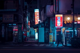 A dimly lit urban street scene featuring neon signs with Korean text. The street is narrow, lined with small shops and businesses, and has a moody, nighttime atmosphere. Bright neon colors illuminate the setting, casting vibrant reflections off the pavement. A person is walking on the sidewalk, adding a sense of life to the quiet street.