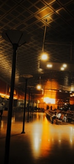 A spacious interior of a dimly lit waiting area featuring an orange glow from large windows at the back. The ceiling has a geometric grid pattern and multiple tall light poles. Rows of empty seats line one side, with a few people seated and scattered in the space. Reflection of light creates a warm ambiance on the polished floor.