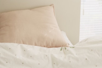 A soft, beige pillow is resting against a creamy white wall, with a light-colored bedspread featuring subtle, diamond-patterned embroidery. A sprig of delicate green plant lies on the bedspread. Bright, natural light filters through a nearby window with white blinds.