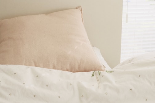A soft, beige pillow is resting against a creamy white wall, with a light-colored bedspread featuring subtle, diamond-patterned embroidery. A sprig of delicate green plant lies on the bedspread. Bright, natural light filters through a nearby window with white blinds.