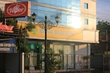 A street view featuring a building with large windows reflecting light. The signage on the building reads 'Bank Bukopin'. In the foreground, a tall street lamp with intricate designs and an orange 'dr. Vapor' sign are visible. Surrounding the building are greenery and banners, one of which mentions COVID-19.