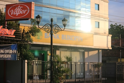 A street view featuring a building with large windows reflecting light. The signage on the building reads 'Bank Bukopin'. In the foreground, a tall street lamp with intricate designs and an orange 'dr. Vapor' sign are visible. Surrounding the building are greenery and banners, one of which mentions COVID-19.