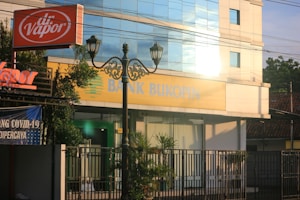 A street view featuring a building with large windows reflecting light. The signage on the building reads 'Bank Bukopin'. In the foreground, a tall street lamp with intricate designs and an orange 'dr. Vapor' sign are visible. Surrounding the building are greenery and banners, one of which mentions COVID-19.