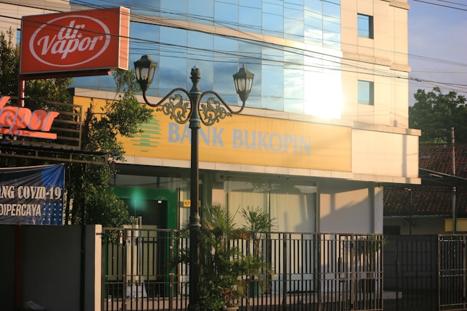 A street view featuring a building with large windows reflecting light. The signage on the building reads 'Bank Bukopin'. In the foreground, a tall street lamp with intricate designs and an orange 'dr. Vapor' sign are visible. Surrounding the building are greenery and banners, one of which mentions COVID-19.