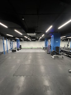 A modern gym with a minimalist interior design, featuring black flooring with interlocking rubber mats. The walls are lined with exercise equipment such as weights and racks. Overhead, evenly spaced ceiling lights provide bright illumination.