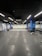 A modern gym with a minimalist interior design, featuring black flooring with interlocking rubber mats. The walls are lined with exercise equipment such as weights and racks. Overhead, evenly spaced ceiling lights provide bright illumination.