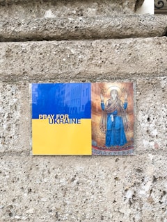 Two images are displayed on a stone wall. The first image on the left has a blue and yellow background with the text 'PRAY FOR UKRAINE'. The second image on the right is a religious icon depicting a haloed figure with hands raised.