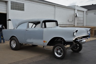 A classic car with a raised front suspension is parked outside a garage. The car has a vintage design with rounded headlights and is unfinished, showing patches of primer paint. Its hood is open, and it rests on shiny chrome wheels. A person is seen near the garage entrance, though only partially visible.