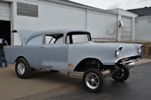 A classic car with a raised front suspension is parked outside a garage. The car has a vintage design with rounded headlights and is unfinished, showing patches of primer paint. Its hood is open, and it rests on shiny chrome wheels. A person is seen near the garage entrance, though only partially visible.