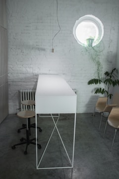 A minimalist interior with white brick walls and a circular window featuring a potted plant. A tall, narrow white table with a metal frame stands next to a wheeled stool. Wooden chairs are placed against the wall, and a pendant light hangs from the ceiling.