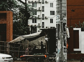 A street scene featuring a small vendor stall situated under a makeshift tent, with shadowy figures inside. The background shows a multi-story building with numerous air conditioning units. A large concrete pole adorned with posters stands prominently in the foreground, next to a white car.